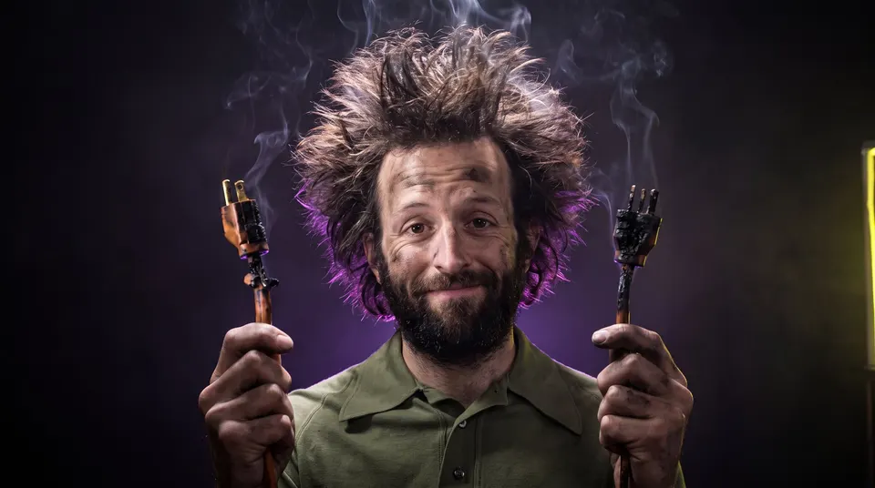 Fried Guy grins sheepishly with soot on his cheeks, holding up two scorched electrical plugs